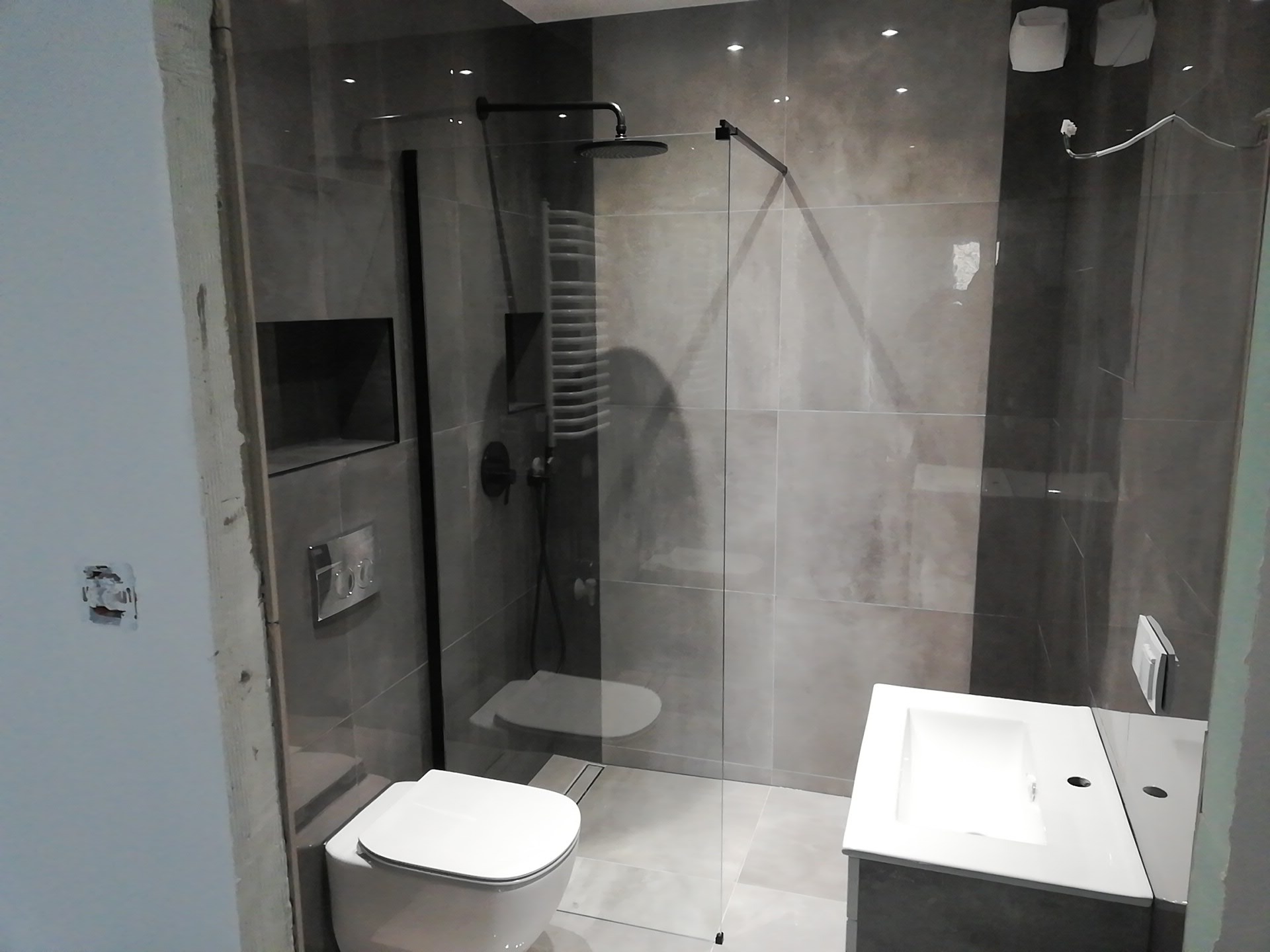 Nowoczesna łazienka z kabiną prysznicową typu walk-in, szare płytki, biała ceramika, czarna armatura i grzejnik drabinkowy, widoczny fragment niedokończonej ściany z instalacją elektryczną.