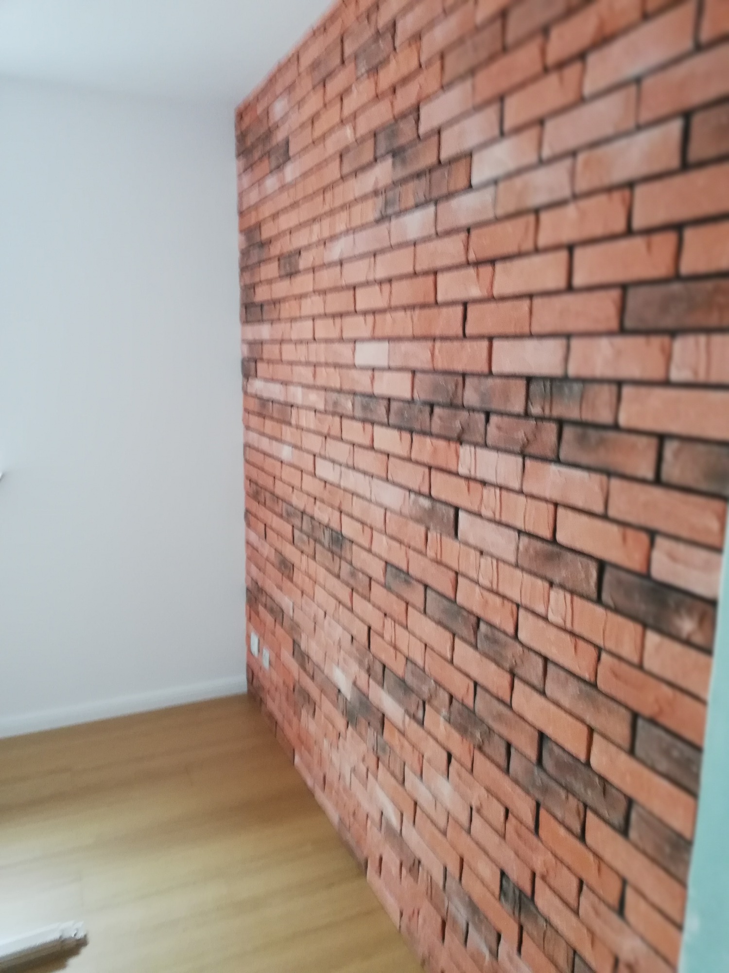 Ściana wyłożona dekoracyjną cegłą w odcieniach czerwieni i brązu, kontrastująca z białą ścianą i jasną podłogą z paneli.