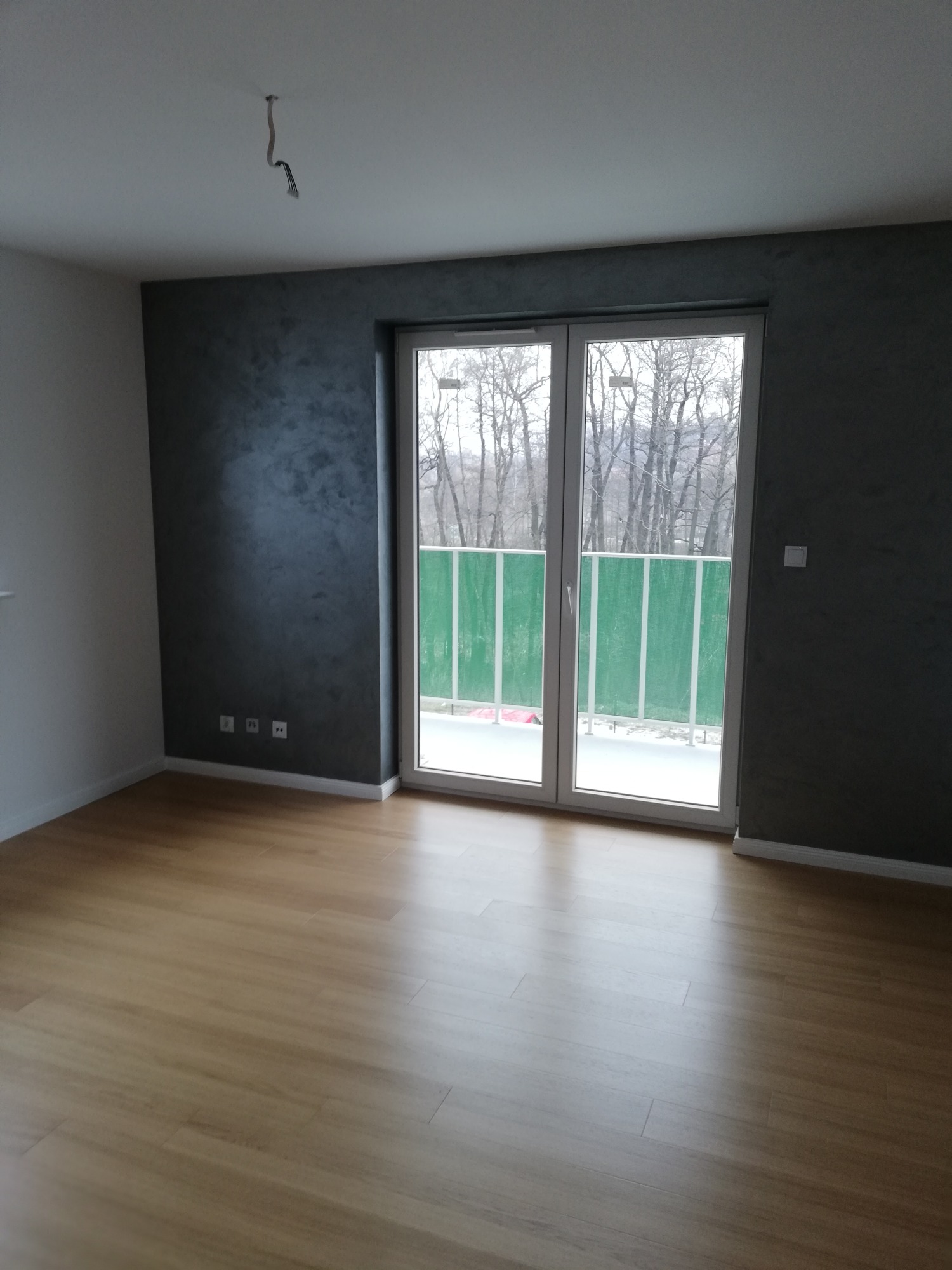 Pusty pokój po remoncie z panelami podłogowymi w odcieniach brązu, ściana pomalowana na ciemny szary z efektem dekoracyjnym, widoczne gniazdka elektryczne i wystające kable z sufitu.