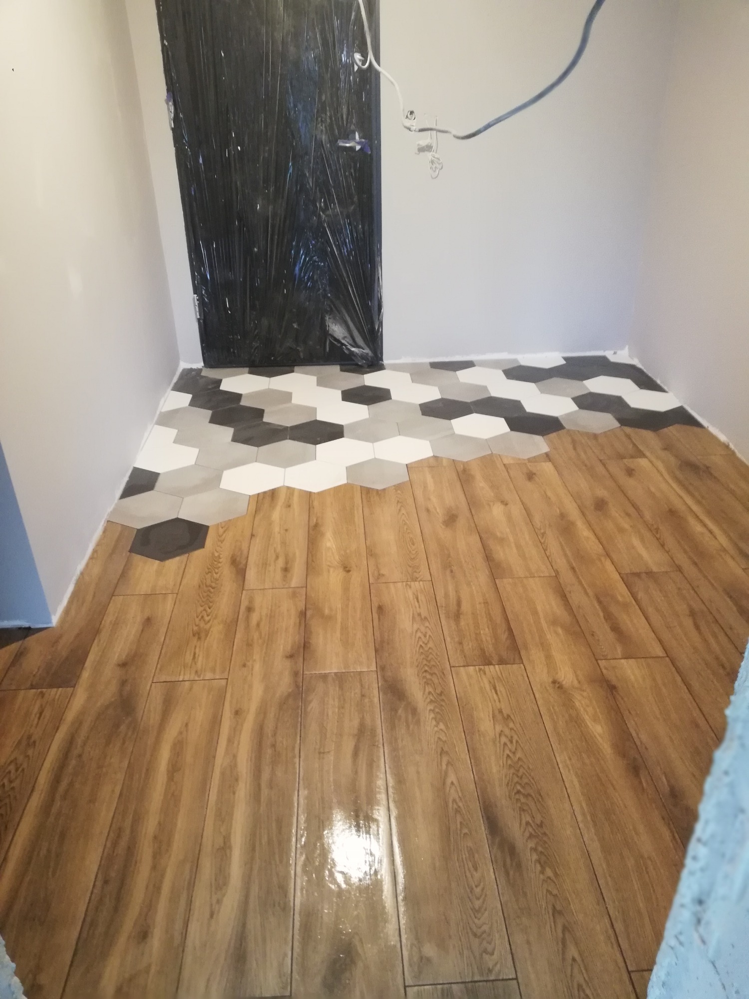 Układanie podłogi: połączenie paneli drewnopodobnych z heksagonalnymi płytkami w odcieniach szarości i bieli. Drzwi zabezpieczone folią ochronną, widoczne przewody elektryczne.