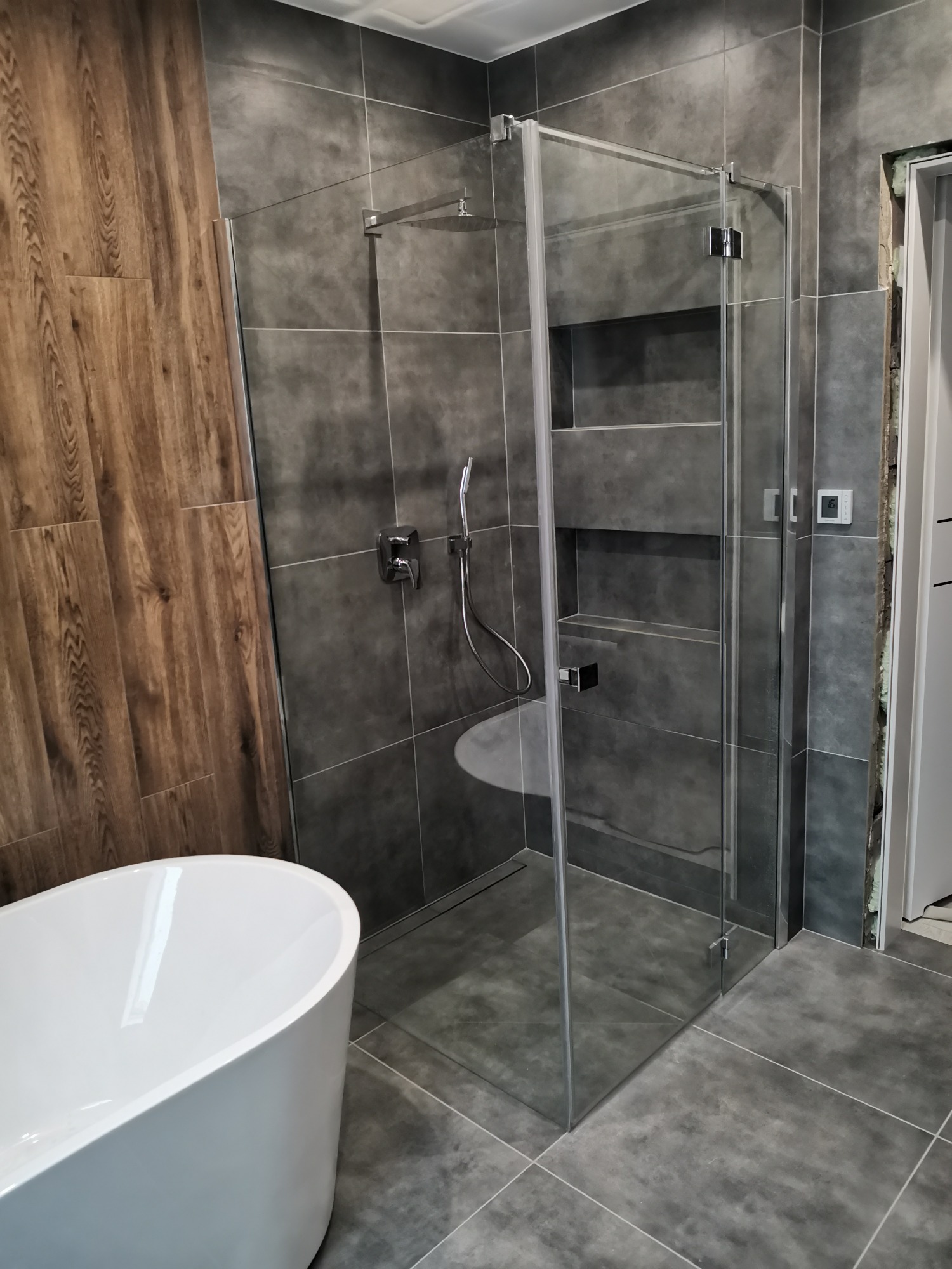 Nowoczesna łazienka z kabiną prysznicową typu walk-in, szare płytki wielkoformatowe, ściana akcentowa z drewnopodobnych paneli, biała wanna wolnostojąca i minimalistyczna armatura.