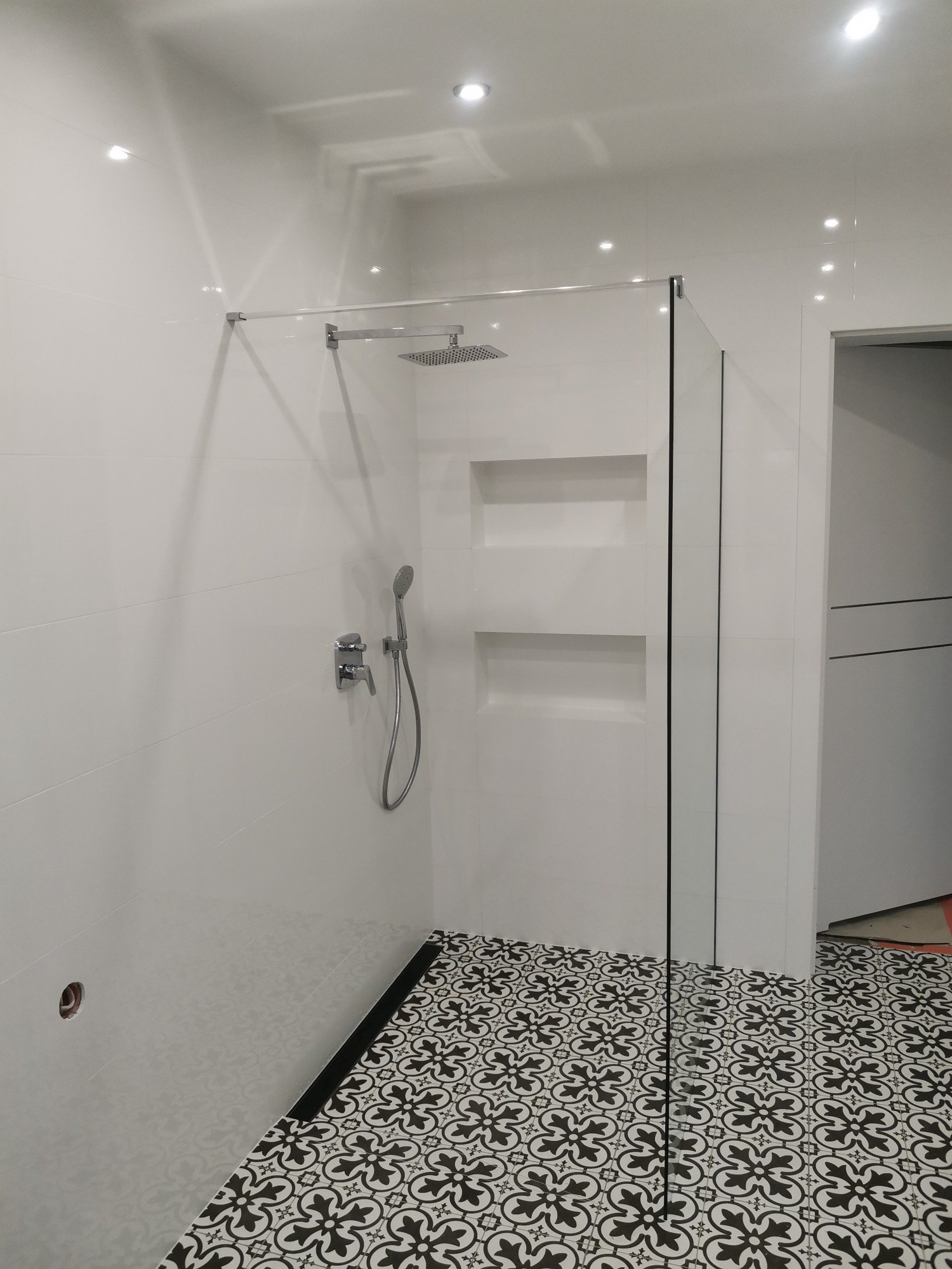 Nowoczesna kabina prysznicowa typu walk-in z czarną ramą, białe płytki ścienne i wzorzysta czarno-biała podłoga, wnęki w ścianie pod prysznicem, minimalistyczna armatura.