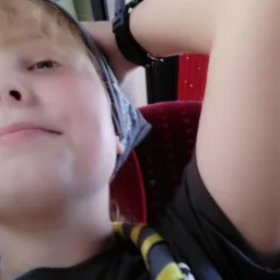 Portret z dołu: blondynka w chuście i koszulce z nadrukiem, siedząca w autobusie z czerwoną zasłoną w tle, z uniesioną ręką w zegarku.