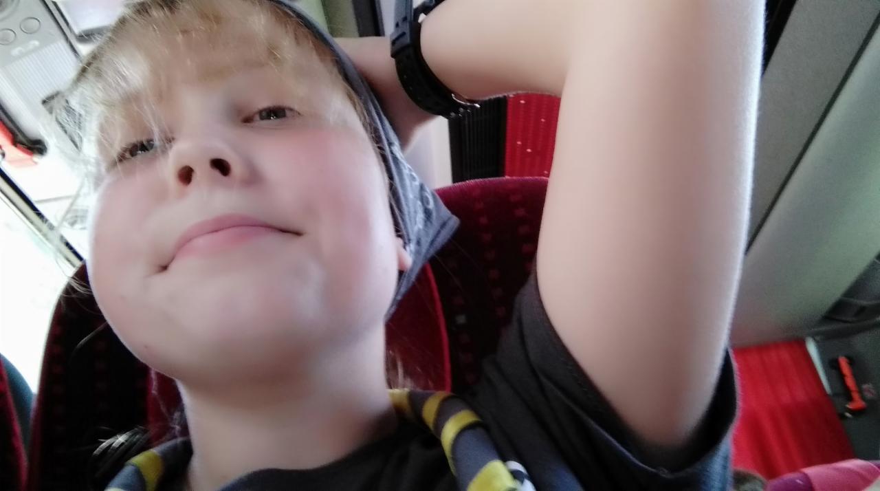 Portret z dołu: blondynka w chuście i koszulce z nadrukiem, siedząca w autobusie z czerwoną zasłoną w tle, z uniesioną ręką w zegarku.