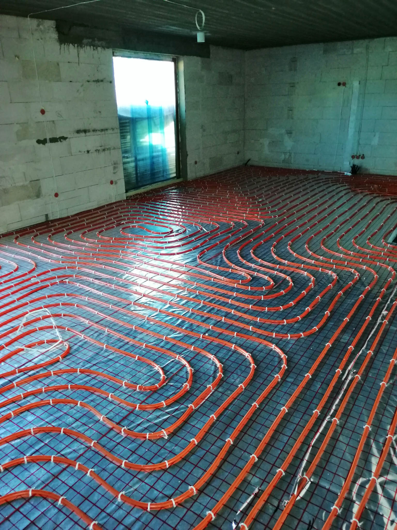 Pomarańczowe rurki ogrzewania podłogowego ułożone na folii izolacyjnej w surowym pomieszczeniu z betonowymi ścianami i otwartym przejściem.