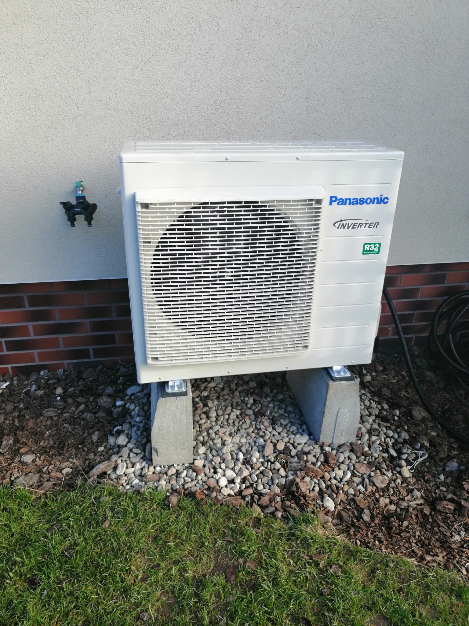 Biała zewnętrzna jednostka klimatyzacji Panasonic Inverter R32, zamontowana na betonowych podstawach, obok ściany budynku z kranem zewnętrznym i elementami ceglanymi.