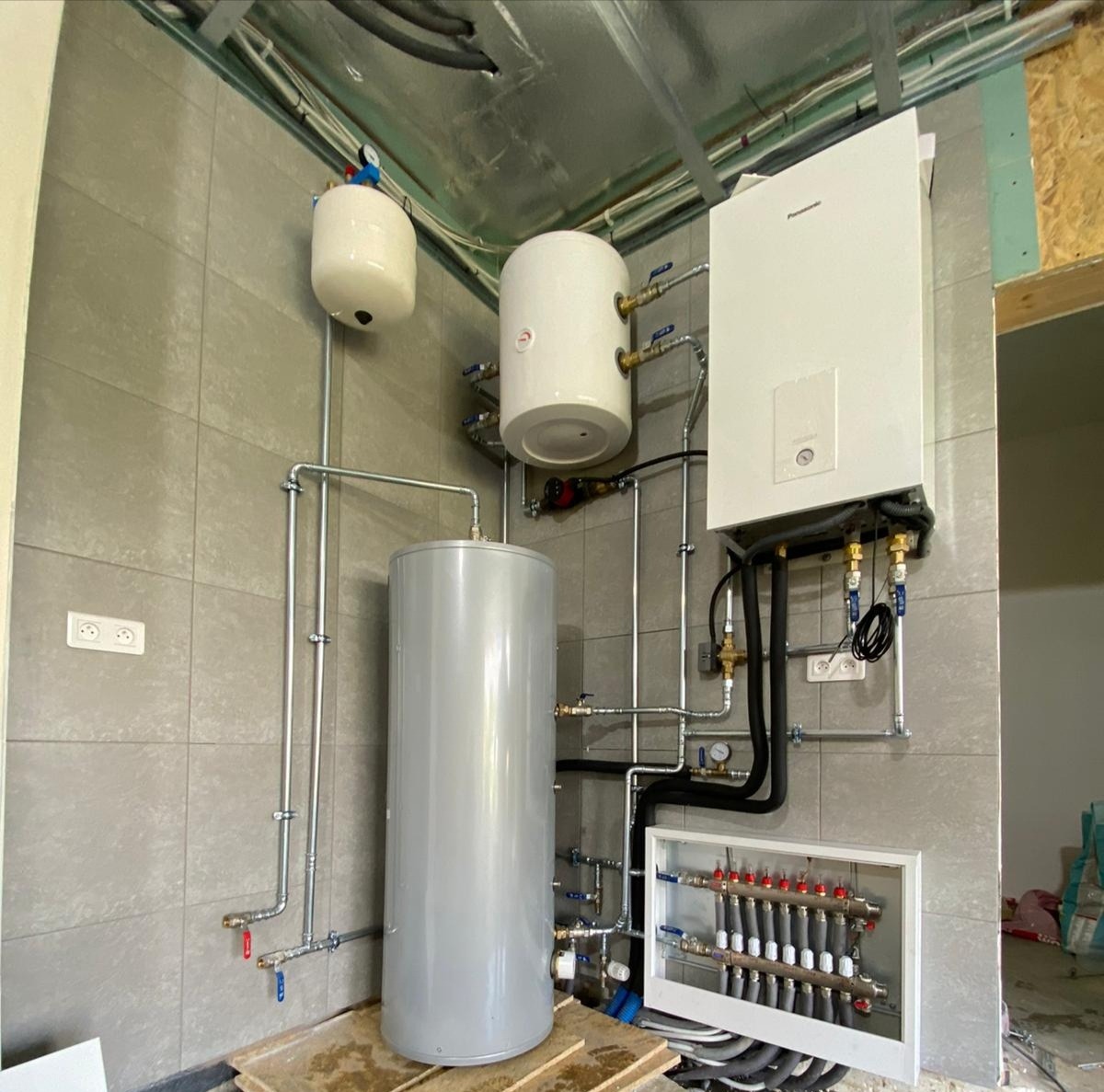 Zmontowana kotłownia w Żelistrzewie: biały piec gazowy Panasonic, szary zbiornik akumulacyjny, białe naczynia wzbiorcze oraz rozdzielacz podłogowy w obudowie, całość połączona rurami na tle szarych...