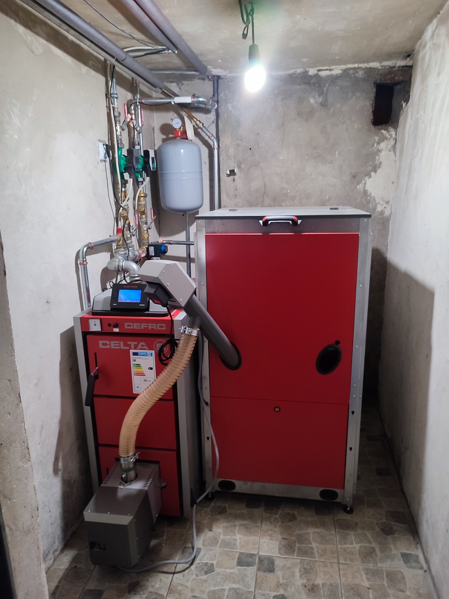 Czerwony piec na ekogroszek Defro Delta z podajnikiem, zainstalowany w piwnicy z widocznymi rurami grzewczymi pod sufitem i surowymi ścianami.