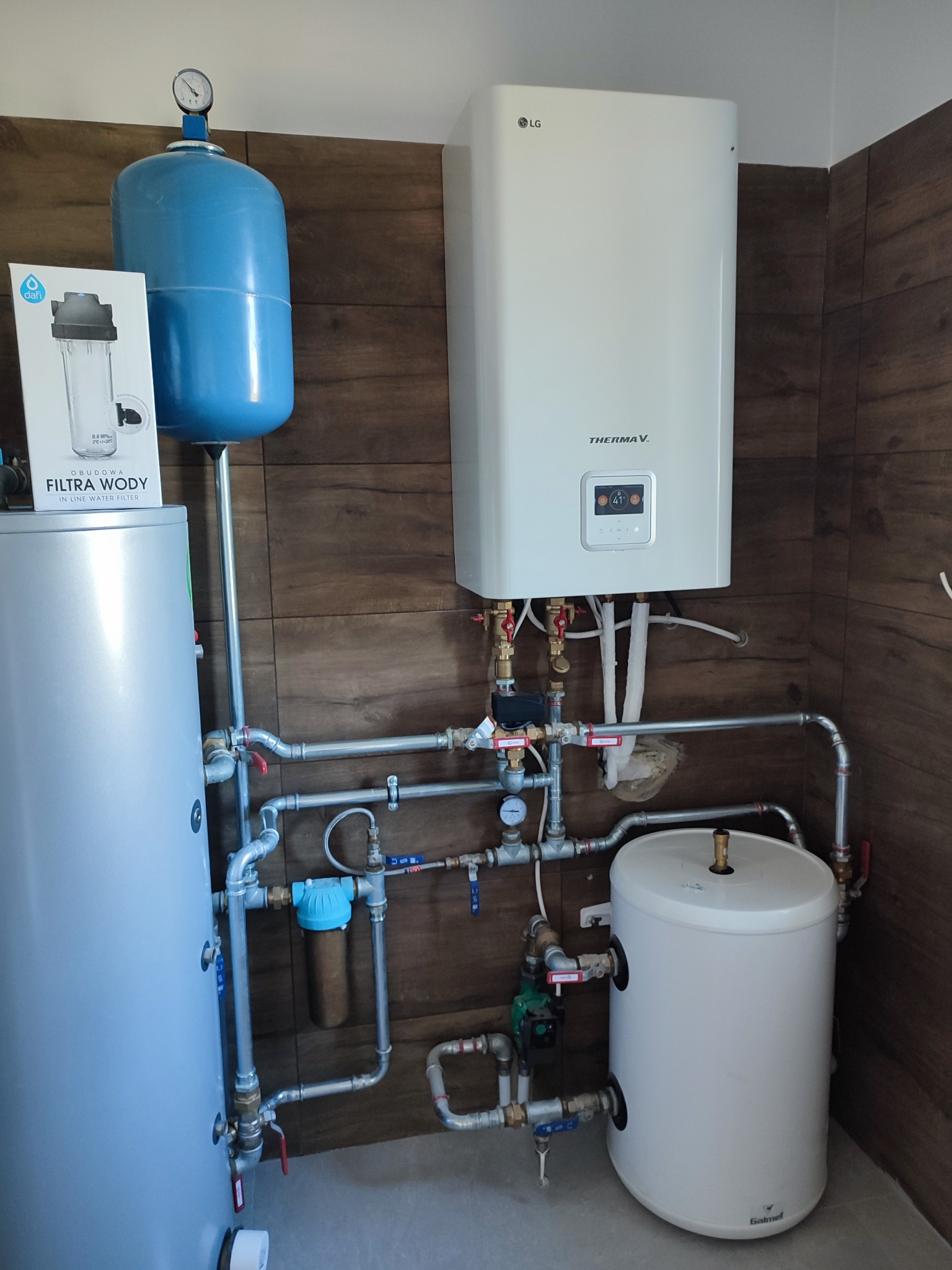 Kompletna instalacja centralnego ogrzewania z pompą ciepła LG Therma V, zbiornikiem buforowym, naczyniem przeponowym i filtrem wody Dafi, zamontowana na tle ściany wyłożonej płytkami imitującymi...