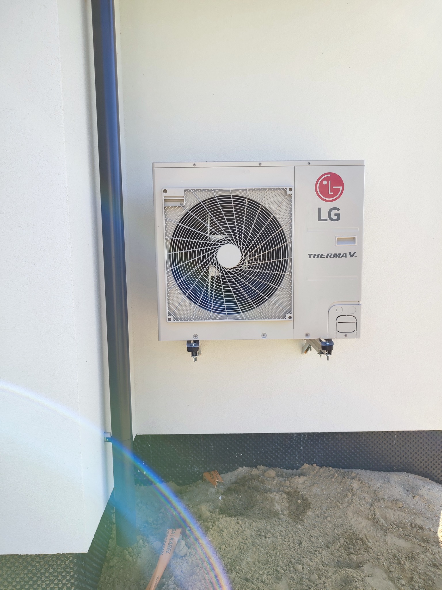 Biała zewnętrzna jednostka pompy ciepła LG Therma V zamontowana na ścianie budynku, widoczne elementy montażowe i rura spustowa, teren wokół przygotowany do dalszych prac ziemnych.