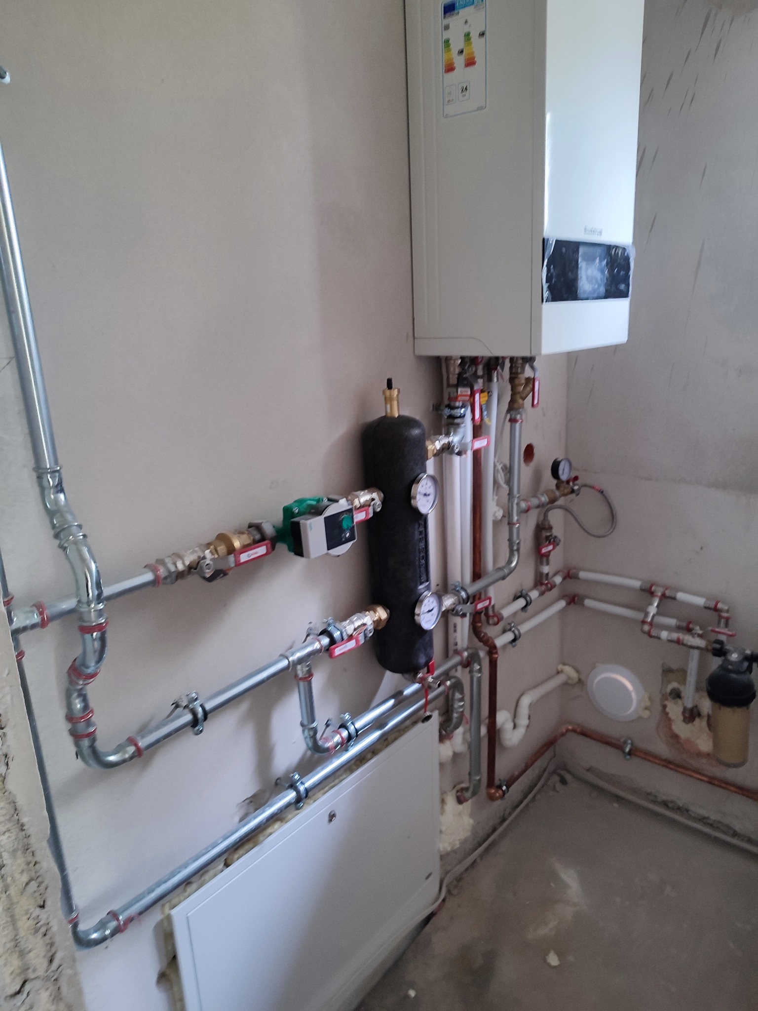 Biały piec gazowy z podłączonymi rurami miedzianymi i stalowymi, czarny zbiornik wyrównawczy z manometrami, widoczne zawory i pompa obiegowa, montaż w surowym pomieszczeniu.