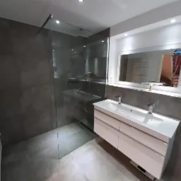 Nowoczesna łazienka z kabiną prysznicową typu walk-in, umywalką nablatową z dwoma kranami i dużym lustrem z oświetleniem LED, wykończona szarymi płytkami imitującymi beton.