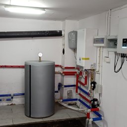 FotonSolar - Kompletna instalacja grzewcza w kotłowni: dwa zbiorniki, piec, inwerter fotowoltaiczny i rozdzielnice. Rury w kolorach czerwonym i niebieskim. Białe ściany i sufit.