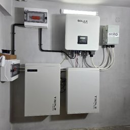 FotonSolar - Kompletny system fotowoltaiczny w pomieszczeniu: dwa akumulatory Triple Power, inwerter Solax, rozdzielnice i okablowanie na szarej ścianie. Gotowa instalacja.