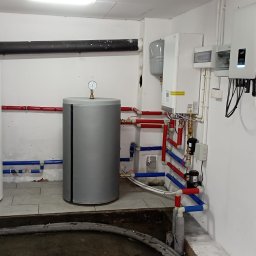 FotonSolar - Instalacja z dwoma zbiornikami, pompą ciepła i rozdzielnicą elektryczną w kotłowni, z widocznymi rurami w kolorach czerwonym i niebieskim.