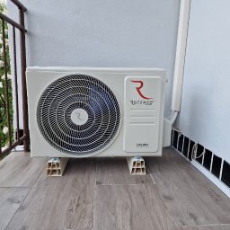 FotonSolar - Biała, zewnętrzna jednostka klimatyzacji Rotenso Ukura, zamontowana na balkonie na plastikowych wspornikach. Widoczne elementy instalacji i kwiaty na balkonie.