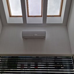 FotonSolar - Biały klimatyzator na ścianie, widok od dołu, trzy okna dachowe z drewnianymi ramami, żaluzje w oknie na dole. Minimalistyczne wnętrze, jasne ściany.