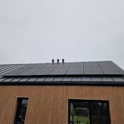 FotonSolar - Dom z panelami fotowoltaicznymi na czarnym dachu z blachy. Widoczne okna i drewniana elewacja. Pochmurne niebo. Ujęcie z dołu.