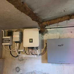 FotonSolar - Instalacja fotowoltaiczna w piwnicy: licznik, inwerter Solax, inwerter FoxESS i bateria. Rury na suficie owinięte jutą. Betonowe ściany. Niski kąt ujęcia.