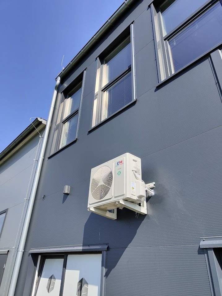 Zewnętrzna jednostka klimatyzacji marki CH zamontowana na elewacji budynku z szarej blachy trapezowej, widoczna instalacja odprowadzająca skropliny.
