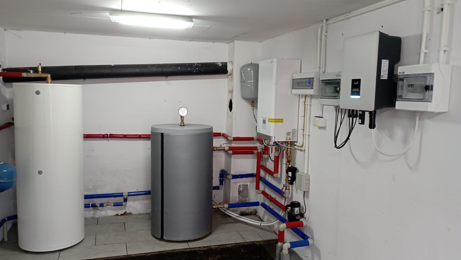 Kompletna instalacja grzewcza w kotłowni: dwa zbiorniki, piec, inwerter fotowoltaiczny i rozdzielnice. Rury w kolorach czerwonym i niebieskim. Białe ściany i sufit.