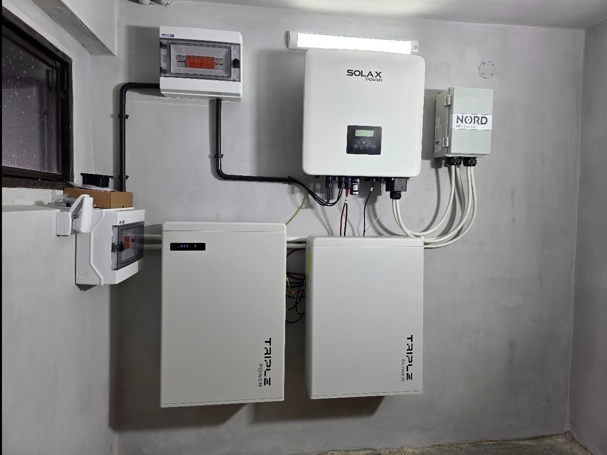 Kompletny system fotowoltaiczny w pomieszczeniu: dwa akumulatory Triple Power, inwerter Solax, rozdzielnice i okablowanie na szarej ścianie. Gotowa instalacja.