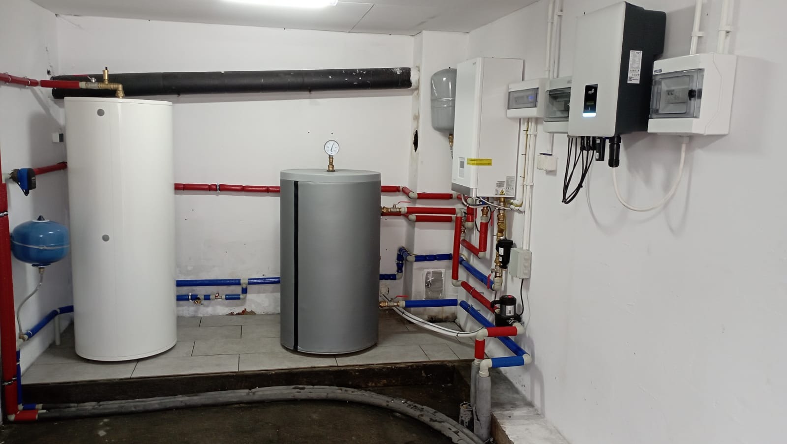 Instalacja z dwoma zbiornikami, pompą ciepła i rozdzielnicą elektryczną w kotłowni, z widocznymi rurami w kolorach czerwonym i niebieskim.