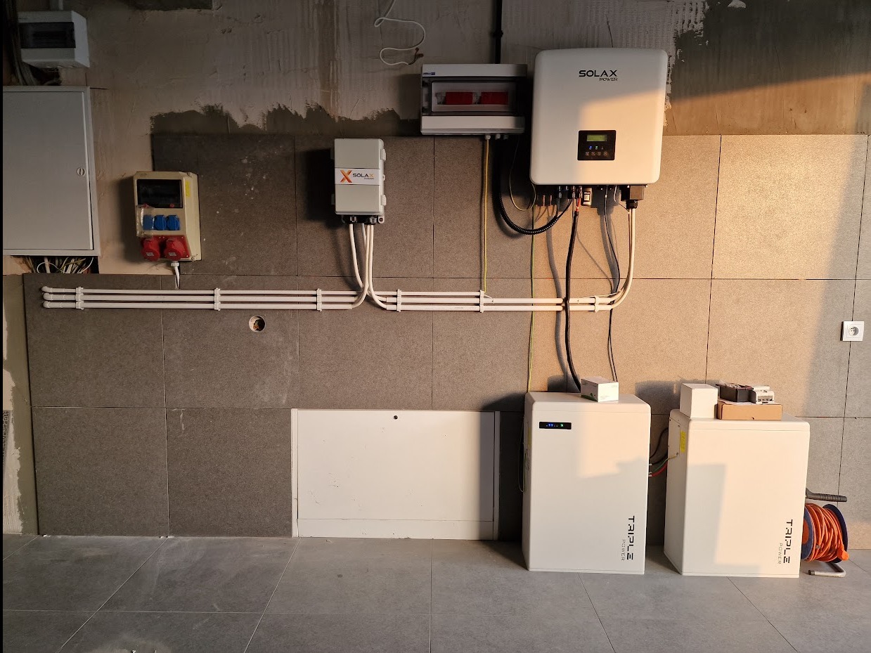 Kompletna instalacja fotowoltaiczna w pomieszczeniu z szarą glazurą: inwerter Solax, magazyny energii Triple Power, okablowanie i rozdzielnice. Widok na ścianę z elementami instalacji.
