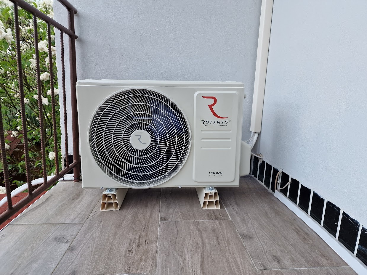 Biała, zewnętrzna jednostka klimatyzacji Rotenso Ukura, zamontowana na balkonie na plastikowych wspornikach. Widoczne elementy instalacji i kwiaty na balkonie.