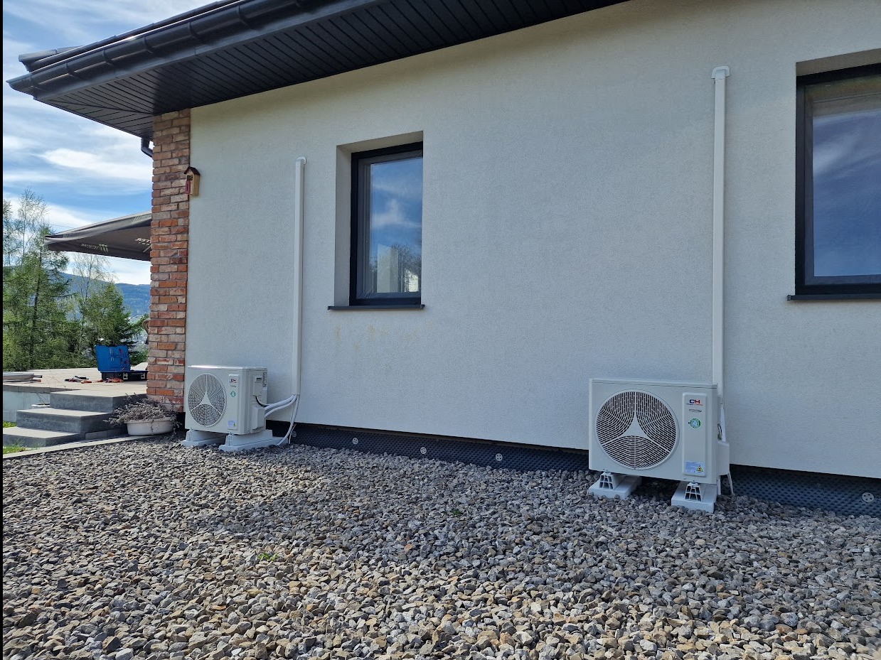 Dwa klimatyzatory zewnętrzne zamontowane na elewacji domu, z widocznymi rurami odprowadzającymi. Tło stanowi elewacja i kamieniste podłoże.