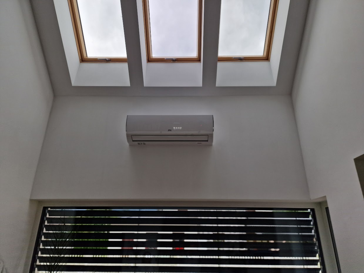 Biały klimatyzator na ścianie, widok od dołu, trzy okna dachowe z drewnianymi ramami, żaluzje w oknie na dole. Minimalistyczne wnętrze, jasne ściany.