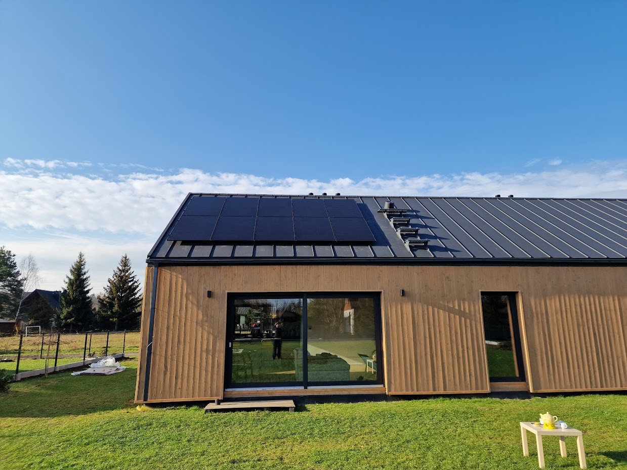 Elegancki dom z panelami słonecznymi na szarym dachu, widok z trawnika z małym stolikiem i serwisem do herbaty. Błękitne niebo z chmurami w tle.