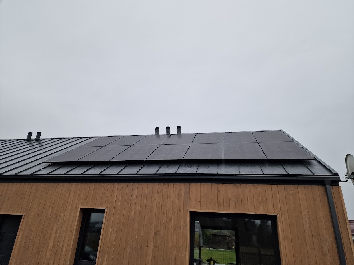 Dom z panelami fotowoltaicznymi na czarnym dachu z blachy. Widoczne okna i drewniana elewacja. Pochmurne niebo. Ujęcie z dołu.