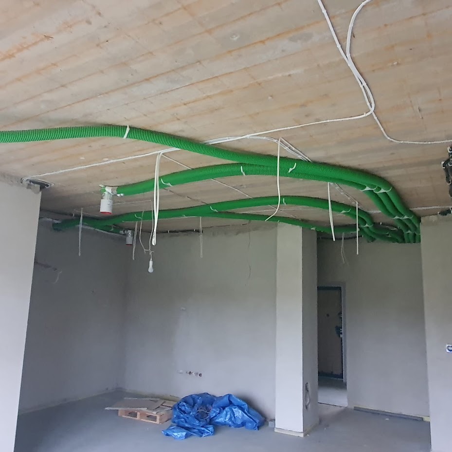 Surowy stan lokalu: zielone, karbowane rury wentylacyjne pod sufitem, białe kable i przewody. Na podłodze folia ochronna i paleta. Widok na szarą ścianę.