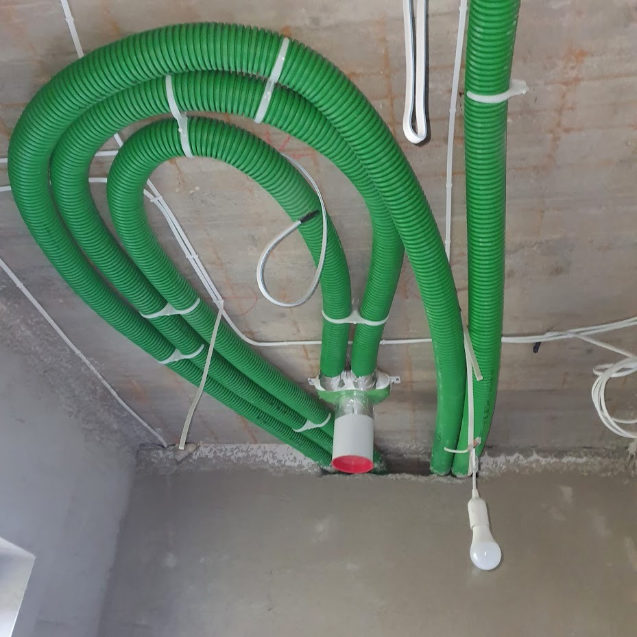 Zielone, karbowane rury wentylacyjne przymocowane do sufitu za pomocą białych opasek zaciskowych. Widoczna żarówka i surowy tynk.