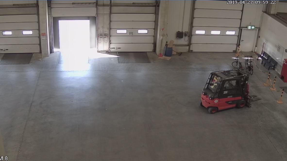 Obraz z kamery przemysłowej w hali magazynowej, widoczny wózek widłowy, rowery i otwarte bramy.