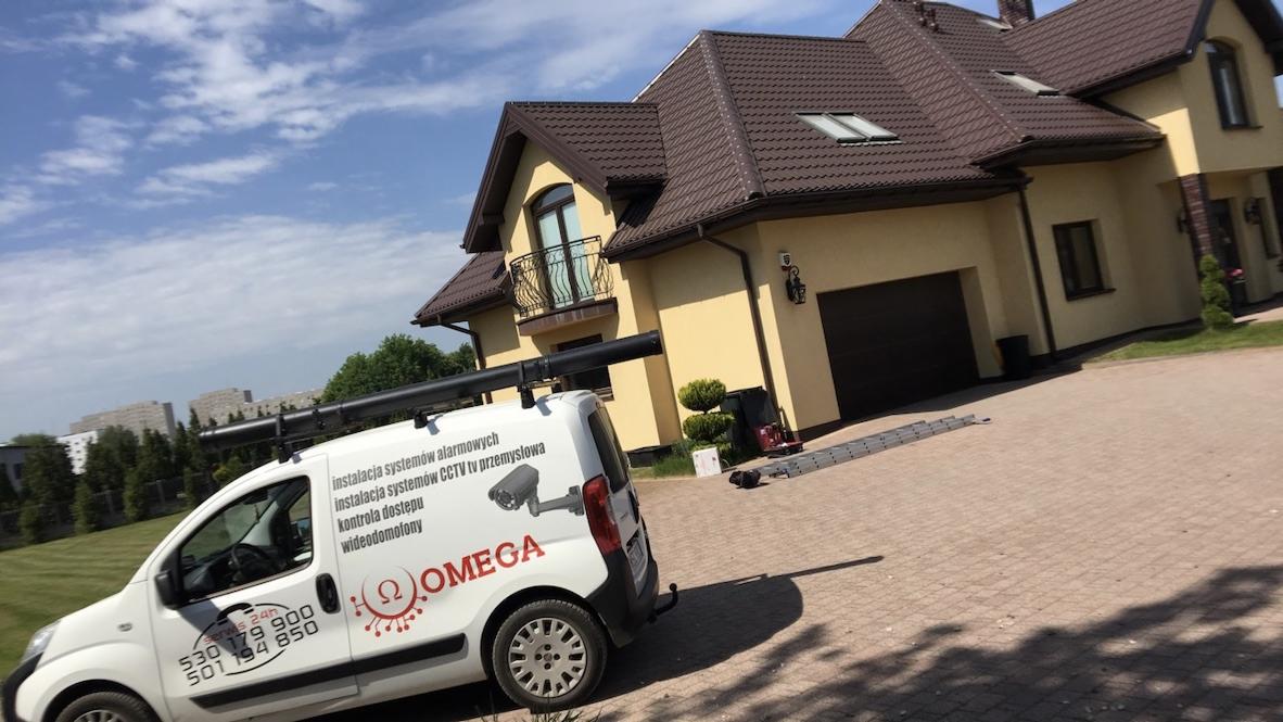 Biały samochód serwisowy z logo firmy Omega, zajmującej się instalacją systemów alarmowych, CCTV i wideodomofonów, zaparkowany przed domem jednorodzinnym z brązowym dachem i beżową elewacją.