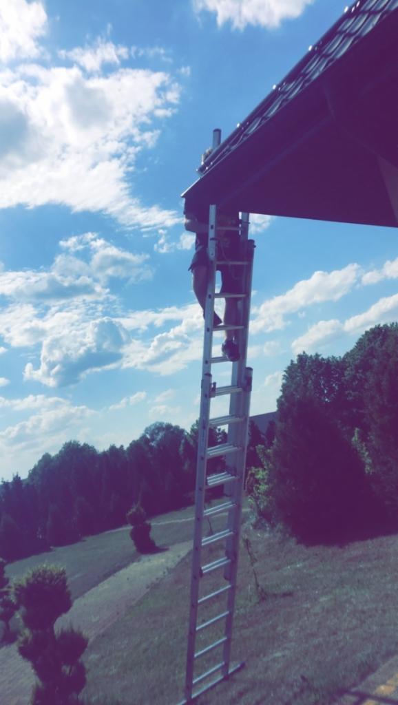 Pracownik na drabinie przy dachu budynku, potencjalnie instalujący system zabezpieczeń lub monitoringu, na tle błękitnego nieba i zieleni drzew.