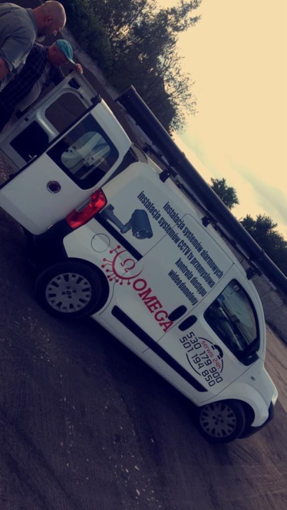 Biały samochód dostawczy z otwartymi drzwiami, oklejony reklamą firmy Omega, specjalizującej się w instalacji systemów alarmowych, CCTV i kontroli dostępu, z widocznymi numerami telefonów...