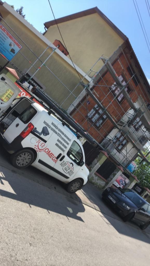 Samochód firmy Omega z logo i numerem telefonu na tle budynku w rusztowaniach, oferującej instalacje systemów alarmowych i CCTV.