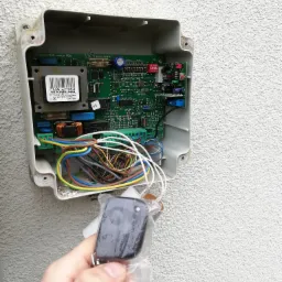 Otwarta obudowa centrali sterującej bramą z widoczną elektroniką, przewodami i ręką trzymającą nowy pilot w plastikowym opakowaniu.