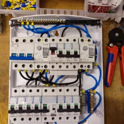 Rozdzielnica elektryczna z zainstalowanymi bezpiecznikami i okablowaniem, widoczne narzędzie do zaciskania końcówek kablowych oraz pudełka z końcówkami kablowymi w różnych kolorach