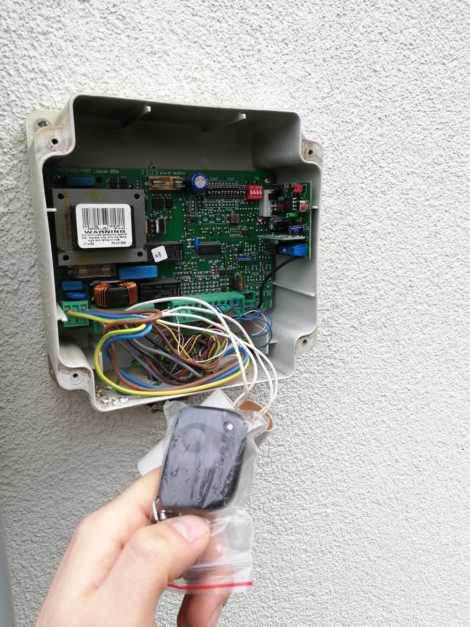 Otwarta obudowa centrali sterującej bramą z widoczną elektroniką, przewodami i ręką trzymającą nowy pilot w plastikowym opakowaniu.