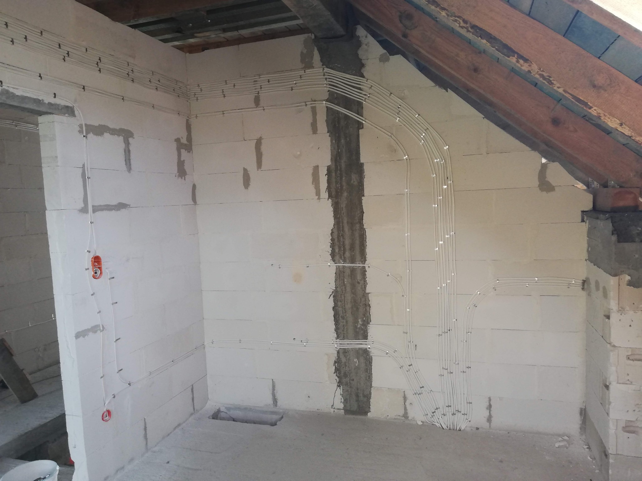 Wnętrze w trakcie prac instalacyjnych: białe przewody elektryczne ułożone równolegle na ścianie z bloczków, mocowane metalowymi uchwytami. Widoczne elementy sufitu oraz fragment otworu drzwiowego...