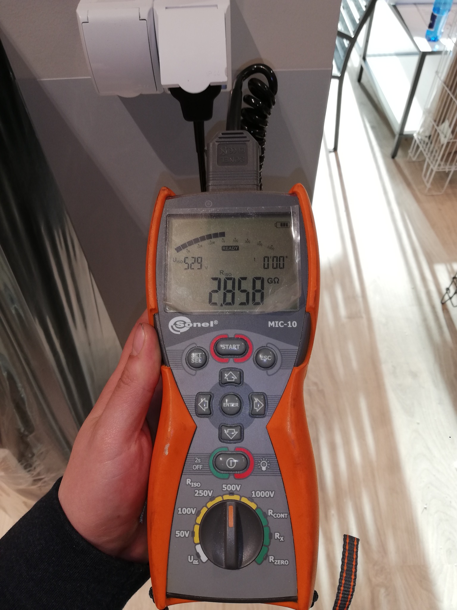 Pomiar rezystancji izolacji gniazdka elektrycznego za pomocą pomarańczowego miernika Sonel MIC-10, wyświetlającego wartość 2.858 GΩ.