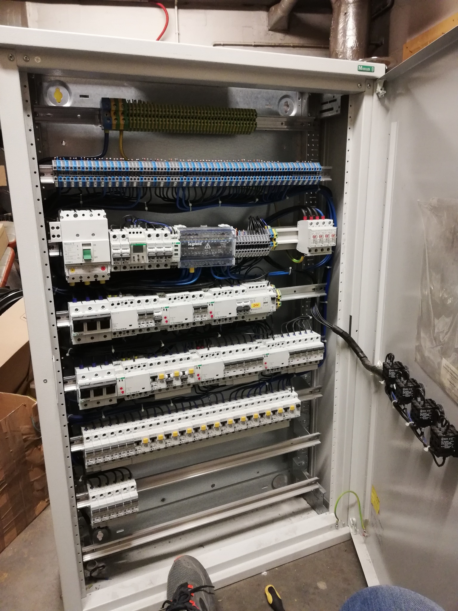 Otwarta szafa sterownicza z zainstalowanymi bezpiecznikami, listwami zaciskowymi i przewodami. Widoczny fragment buta i śrubokręta na podłodze.