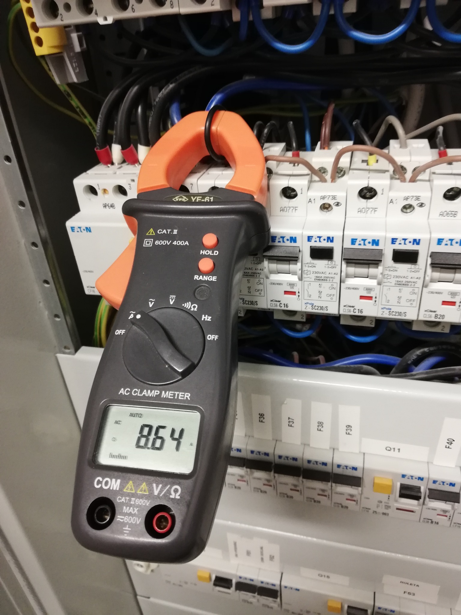 Miernik cęgowy prądu zmiennego YF-61, wskazujący wartość 8.64, zawieszony na przewodach w rozdzielnicy elektrycznej z bezpiecznikami Eaton, widoczne oznaczenia bezpieczników i przewodów.