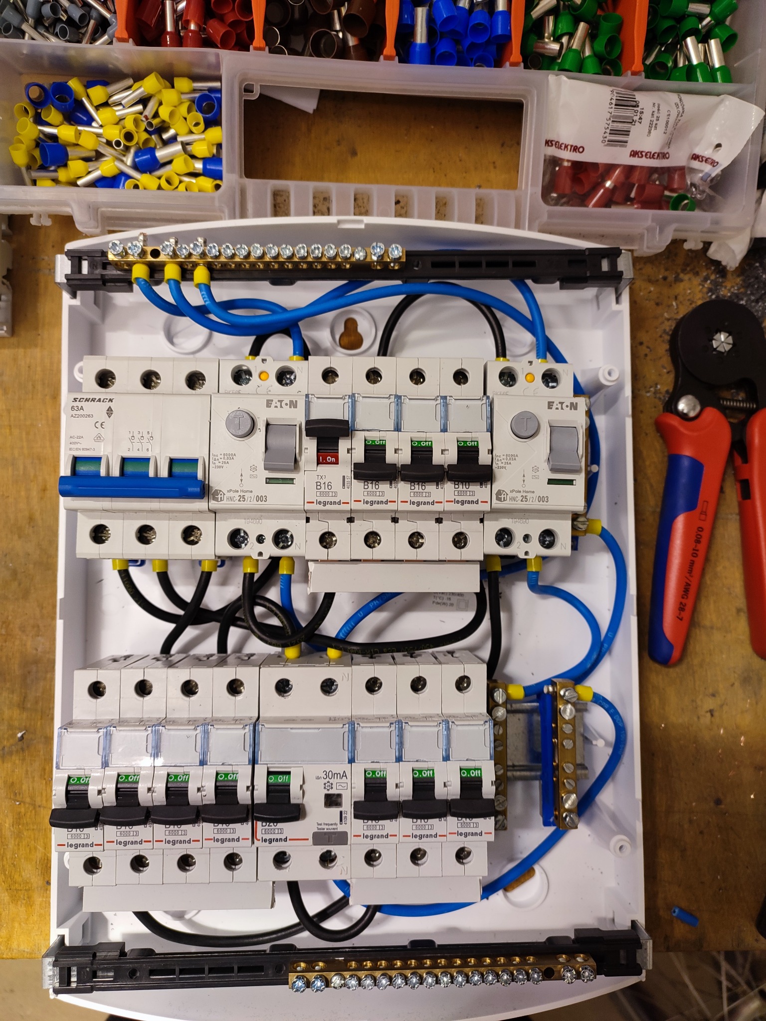 Rozdzielnica elektryczna z zainstalowanymi bezpiecznikami i okablowaniem, widoczne narzędzie do zaciskania końcówek kablowych oraz pudełka z końcówkami kablowymi w różnych kolorach