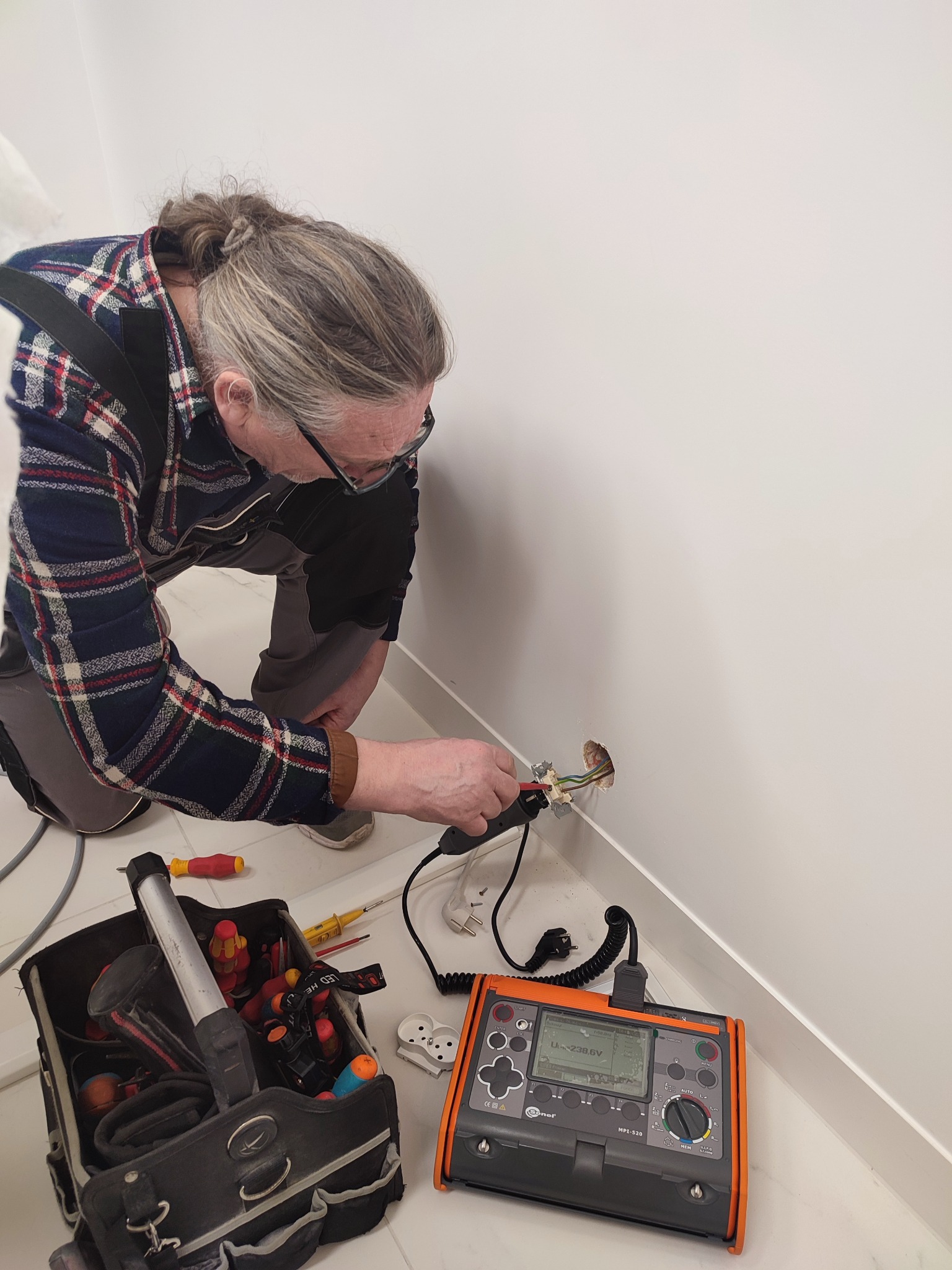 Elektryk w koszuli w kratę, w okularach, wykonuje pomiary instalacji elektrycznej w białym pomieszczeniu za pomocą miernika Sonel MPI-520; obok torba z narzędziami.