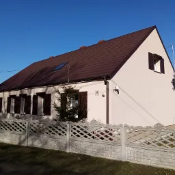 Jasny dom jednorodzinny z brązowym dachem i drewnianymi okiennicami, widoczny numer domu '15' i ozdobny betonowy płot w słoneczny dzień.